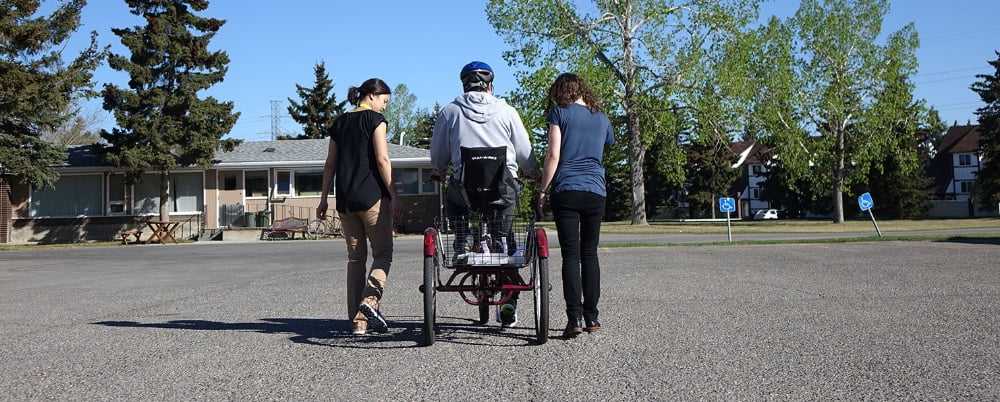 3 people outside, one on each side of an individual that is using a tricycle.