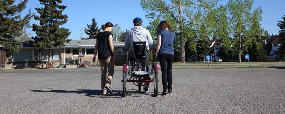 3 people outside, one on each side of an individual that is using a tricycle.